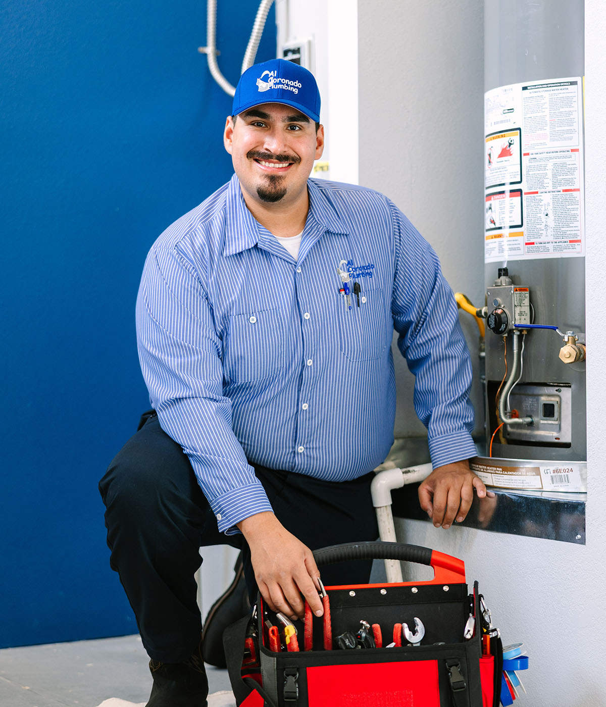 Al Coronado plumber smiling with a toolbox in front of a water heater